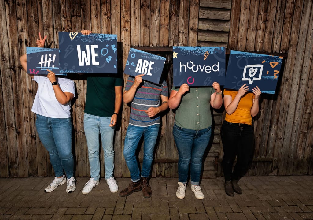 Fünf Personen halten Schilder mit der Aufschrift „Hi! We Are Hooved“ und stehen vor einer Holzwand. Die Gesichter sind von den Schildern verdeckt.